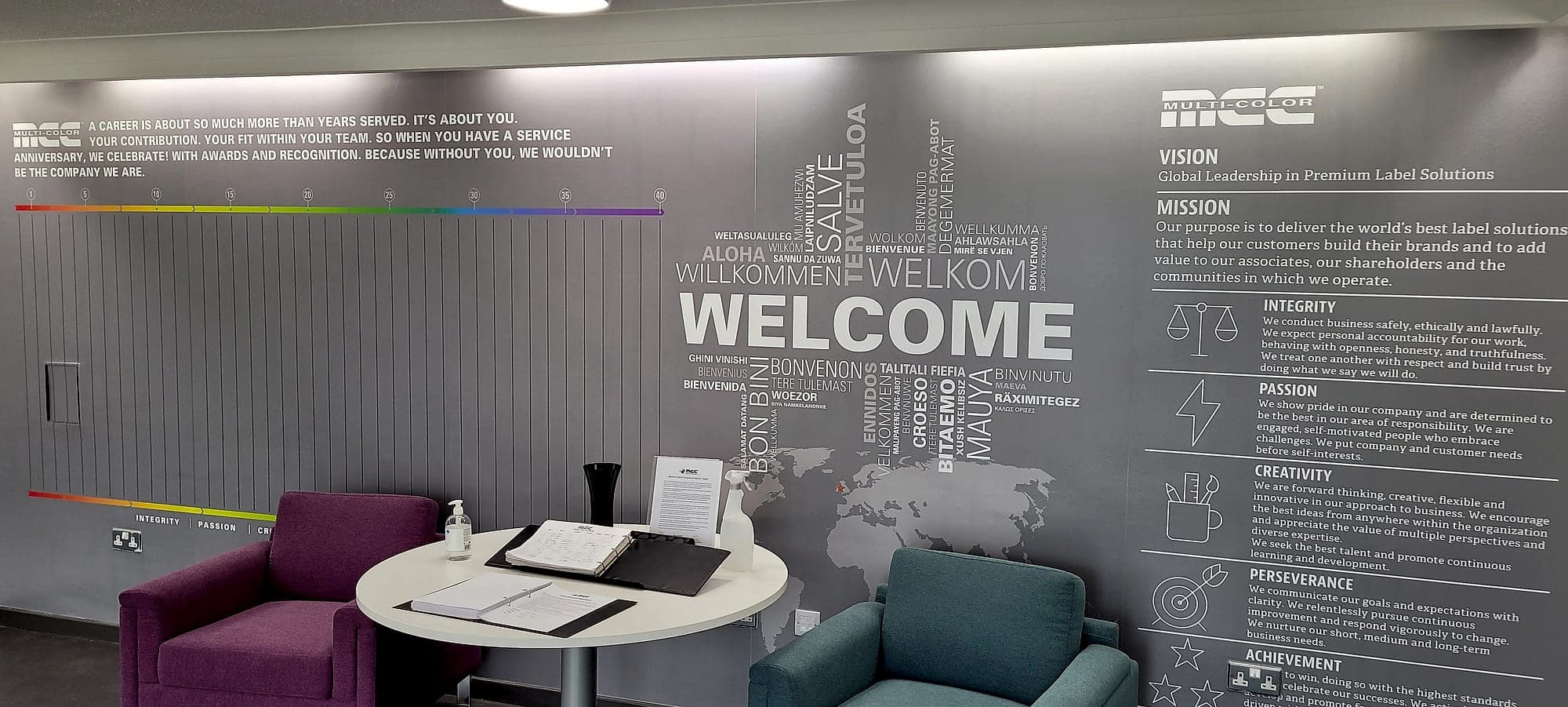 Environmental graphics in a corporate office: a full-height printed wall carrying a welcome in many languages, a service-anniversary timeline and the company's vision, mission and values, with breakout seating in front of it