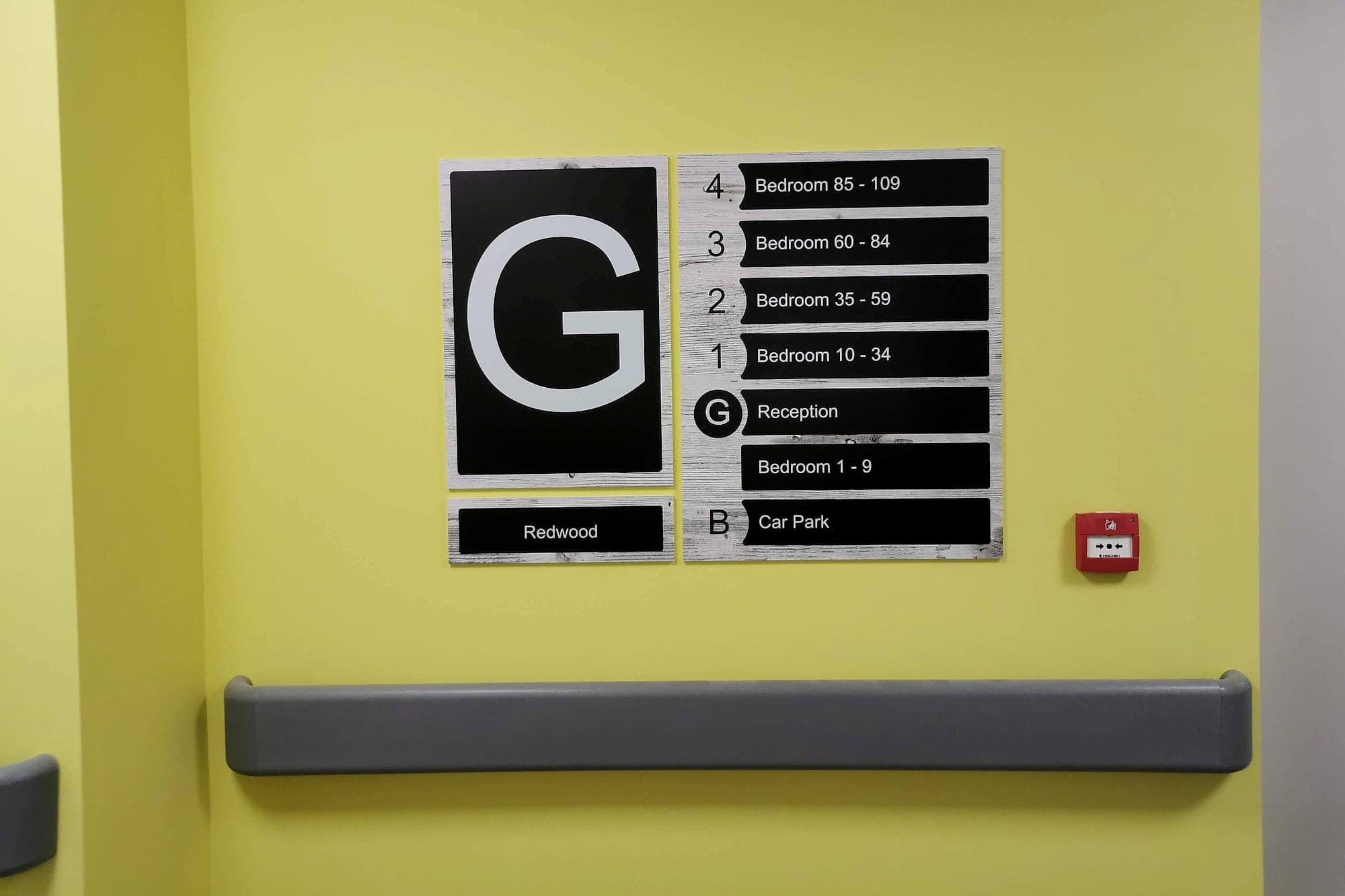 A care-home lift lobby (accessible wayfinding surfaces in a care home): matt black level directory and floor letter on a strong yellow wall above a grey handrail