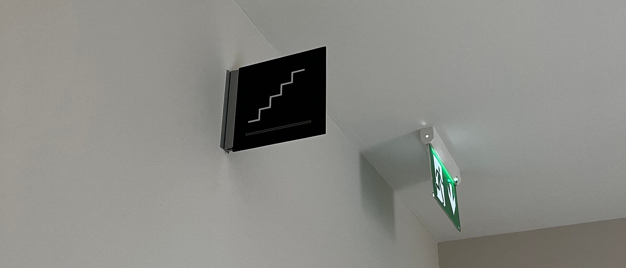 Photoluminescent signage context: a black projecting stair pictogram sign beside a green illuminated running-man emergency exit luminaire at ceiling level in an apartment lobby