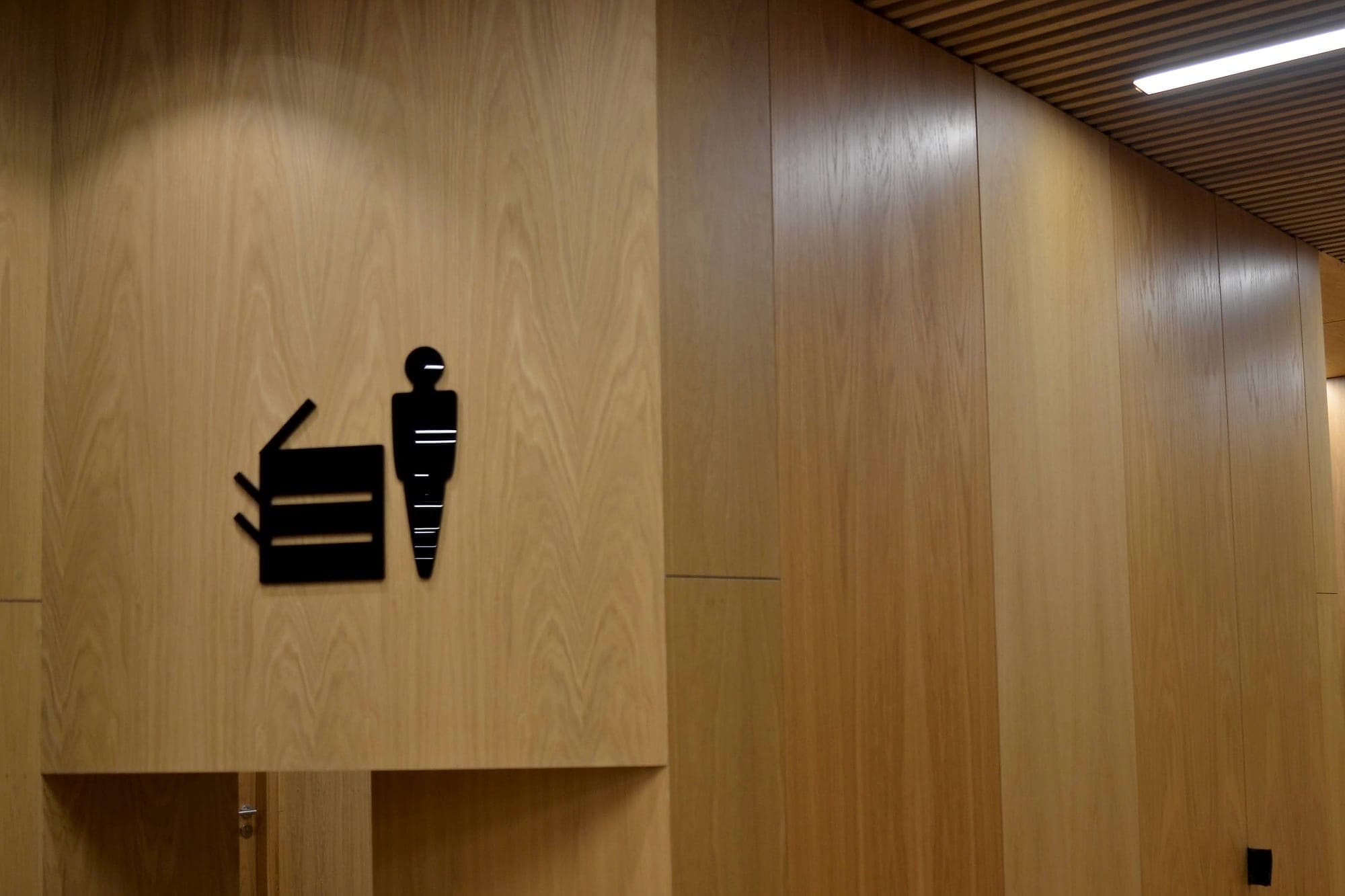 Flush-cut black accessible WC pictogram applied to full-height oak wall panelling at Trinity GBHI, in a contemporary academic interior