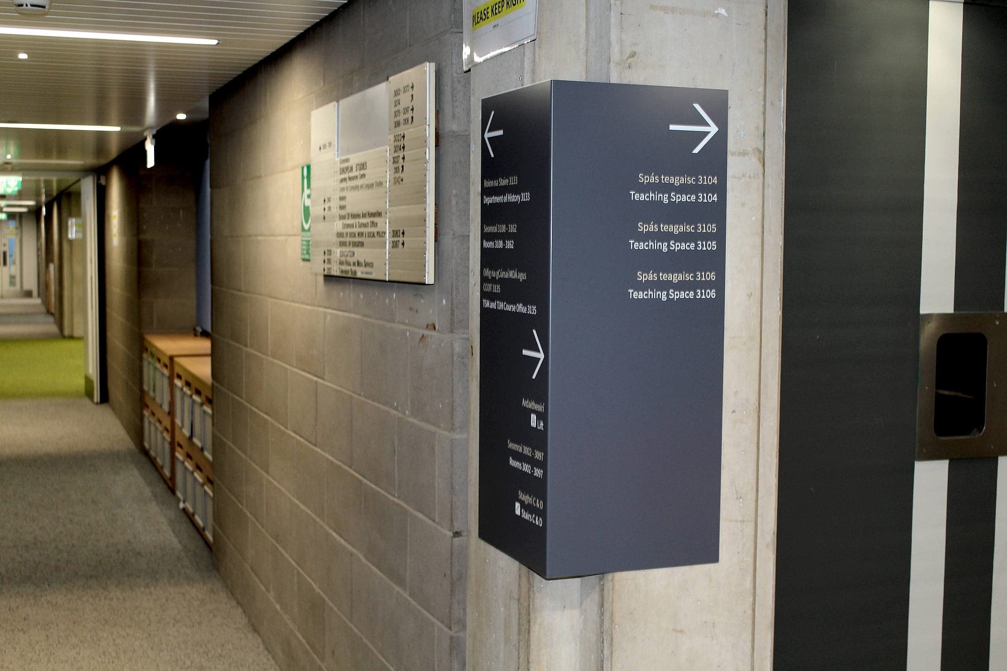 The Arts Block: double-sided projecting sign at a corridor junction listing bilingual room-number ranges, teaching spaces, lift and stairs with left and right arrows