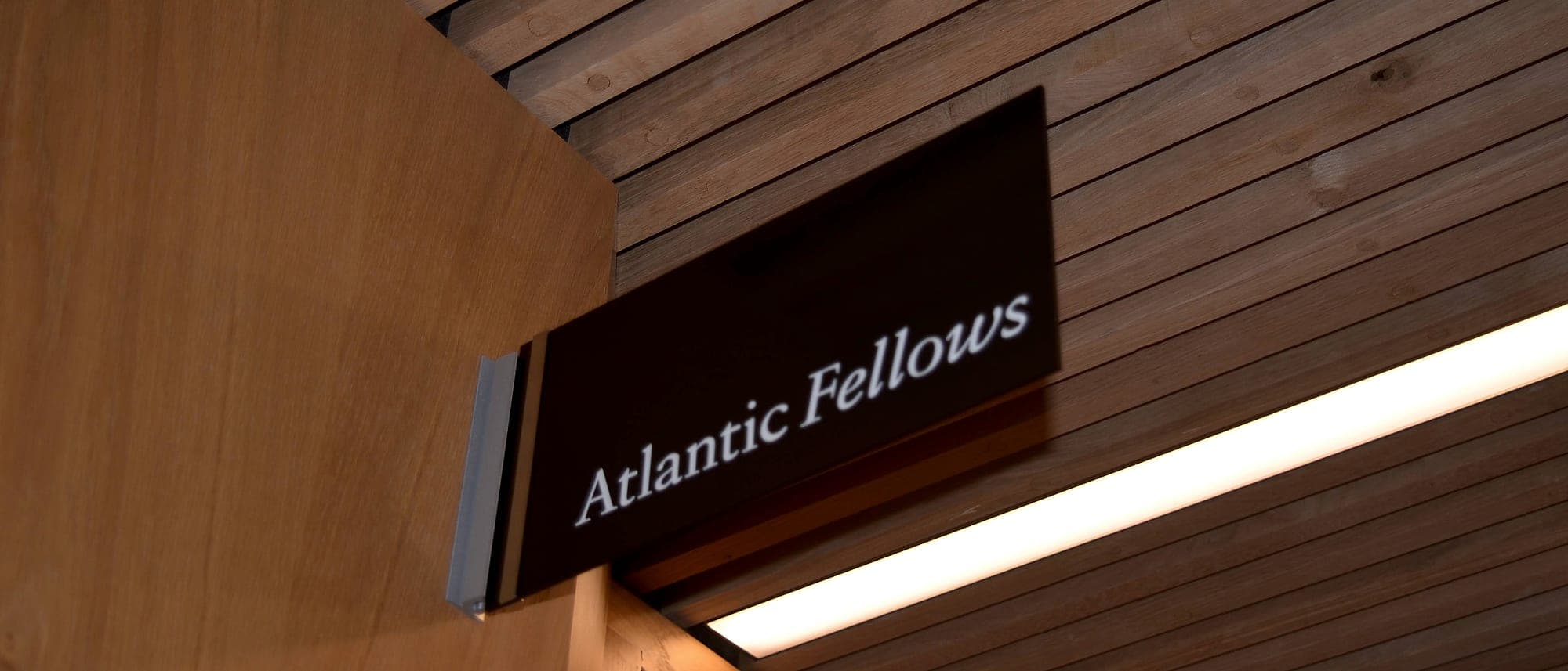 Projecting sign at Trinity GBHI reading Atlantic Fellows, mounted perpendicular to the wall at a timber-lined corridor corner, positioned to be read along the approach