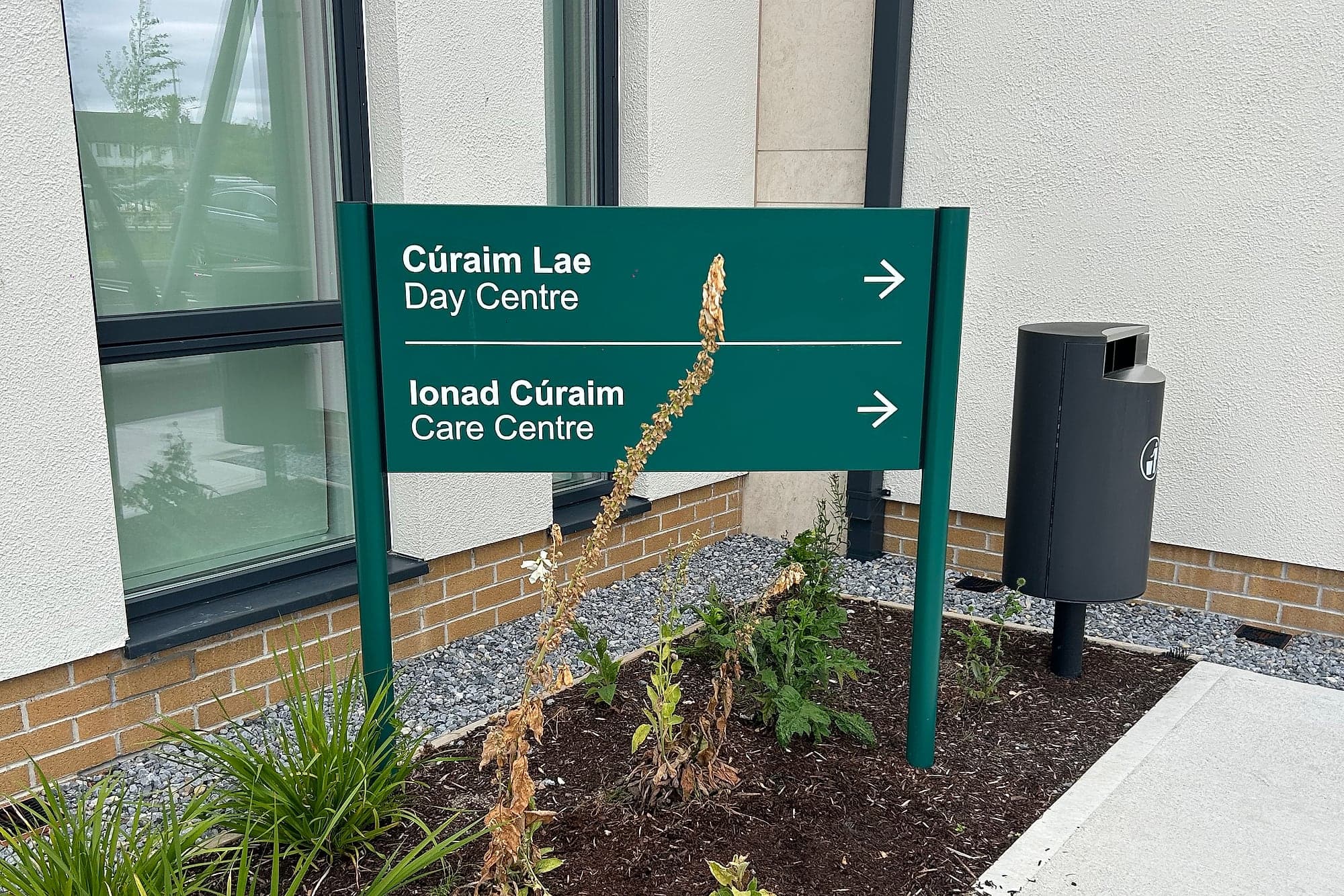 Bilingual Irish and English external post-and-panel directional sign at a community nursing unit, reading Cúraim Lae / Day Centre and Ionad Cúraim / Care Centre, with arrows, in a planted bed