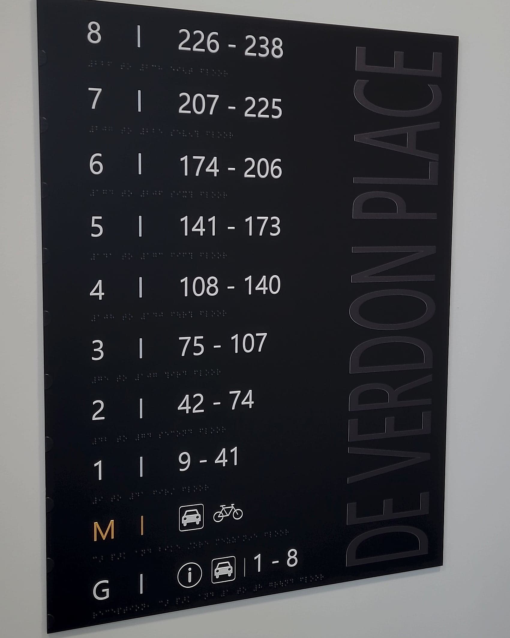 Residential level directory sign at De Verdon Place in black, with floor bands from ground to level 8, apartment number ranges, braille beneath every line and car, bicycle and information pictograms