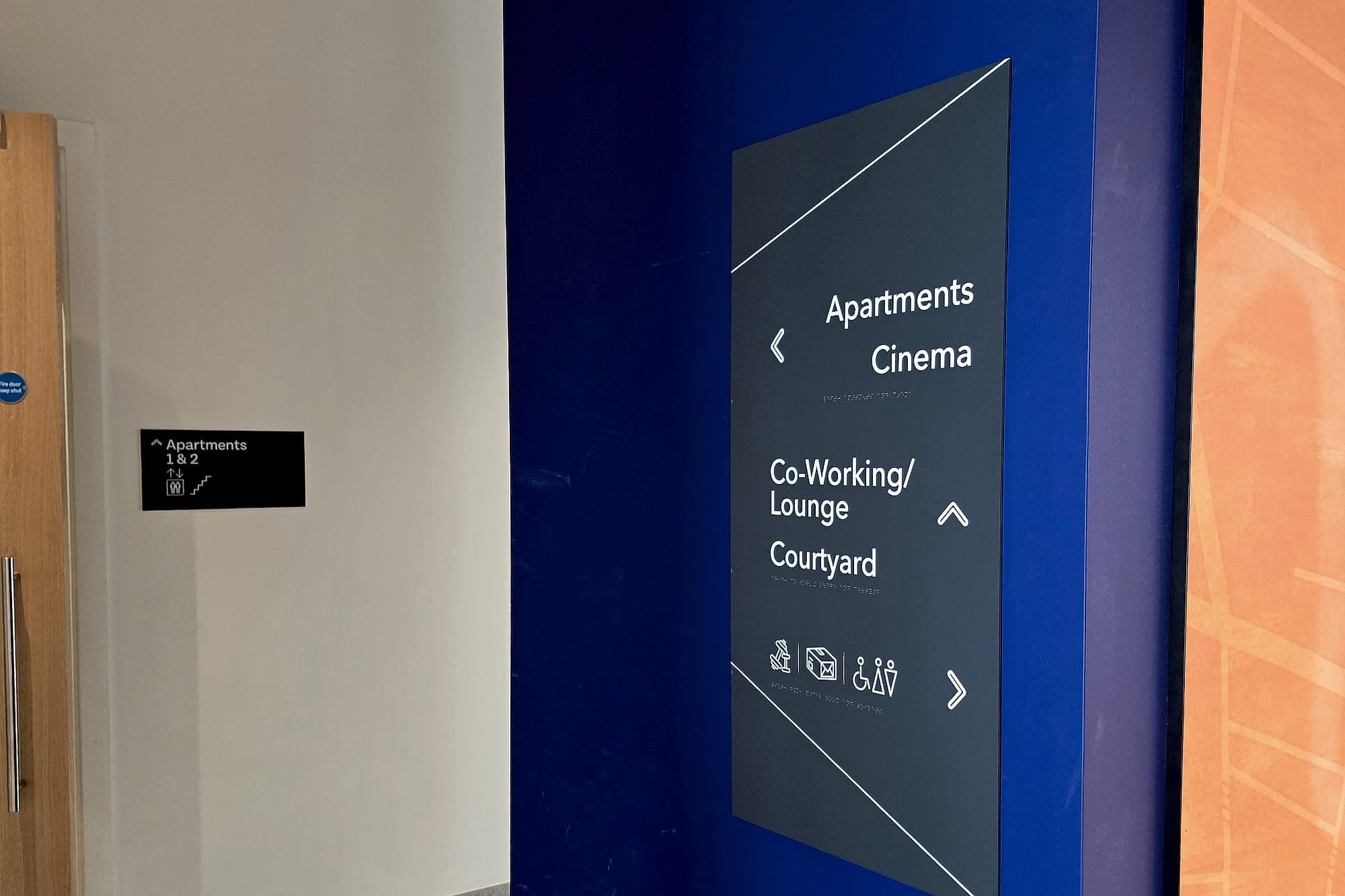 Residential amenity directional signage at the Glass Bottle Quarter listing apartments, cinema, co-working lounge and courtyard, with braille and pictograms, beside a printed street-map wall graphic