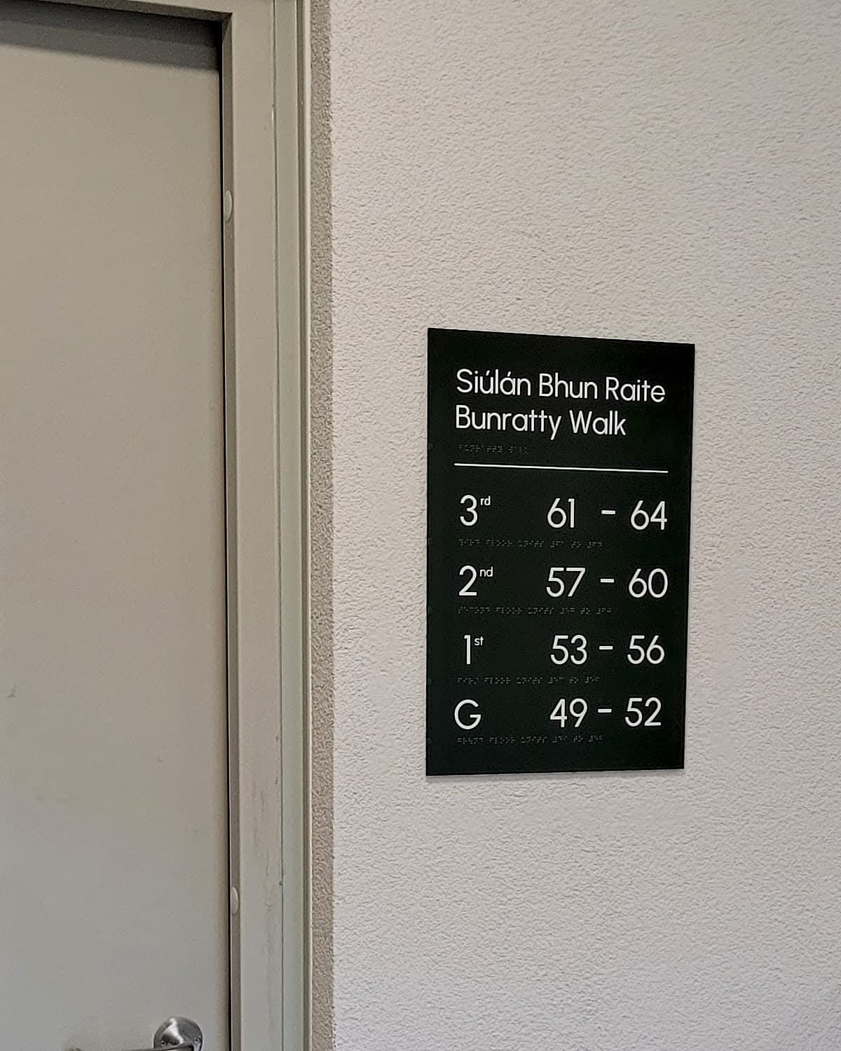 Bilingual Irish and English residential core directory at Bunratty Walk listing each floor against its apartment number range, with braille beneath every line, mounted beside a stair-core door