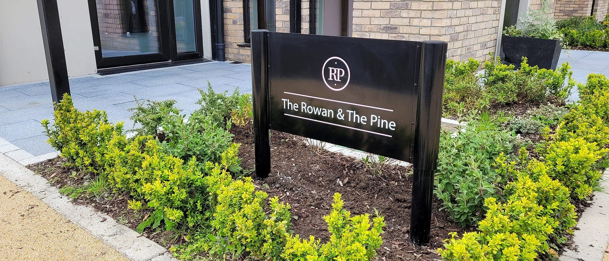 Rathyvoy: external residential entrance signage on a post-and-panel frame set in finished planting outside a newly completed apartment building