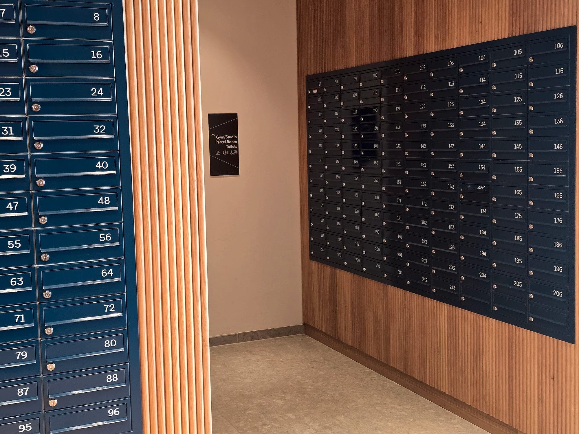 Residential lobby at the Glass Bottle Quarter for Lioncor: two banks of numbered apartment mailboxes on timber-clad walls and a small internal wall sign directing residents to the gym, studio, parcel room and toilets