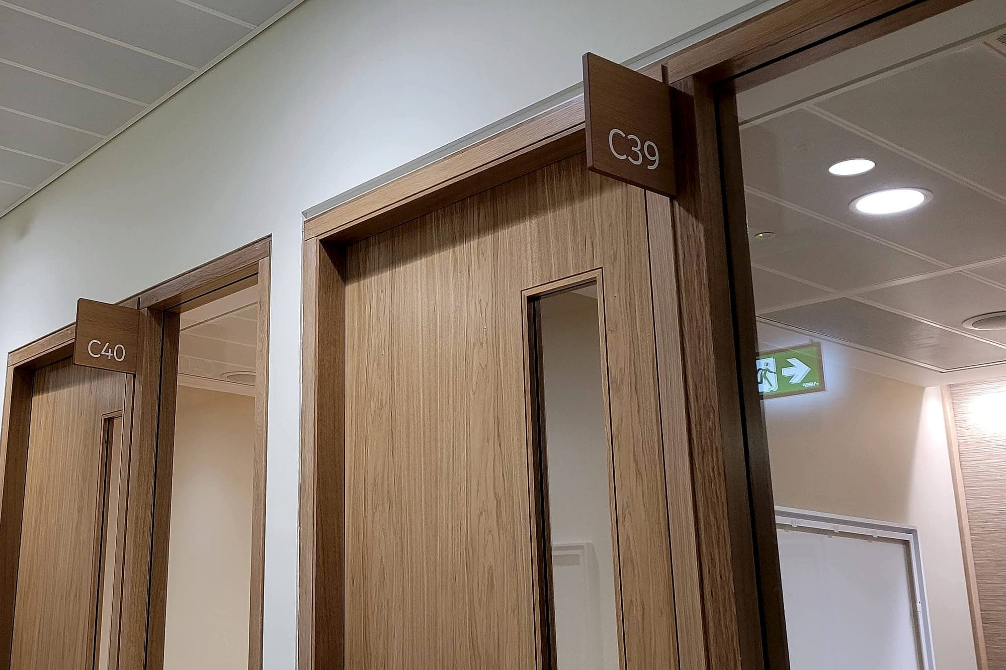 Salesforce: timber projecting room identification signs C39 and C40 over corridor doors, with an internally illuminated green running-man escape sign visible beyond. Safety signage and wayfinding kept in separate height bands
