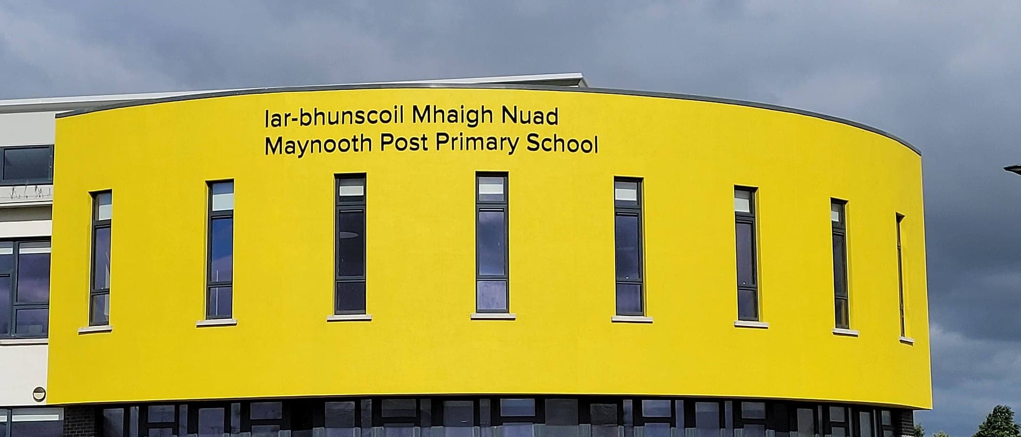 Curved bright yellow facade of Maynooth Post Primary School carrying its bilingual name in black lettering, the building's form and colour doing the arrival wayfinding