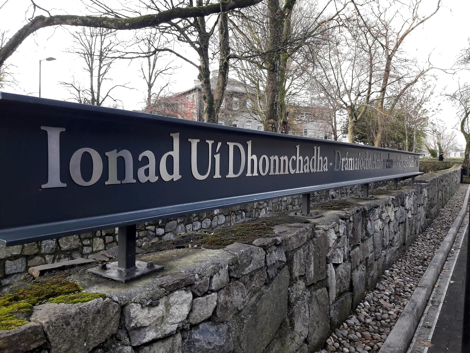 Structural signage installation at the University of Galway: a long dark steel identification sign carrying polished cut lettering, carried on steel posts with bolted baseplates fixed into a stone boundary wall