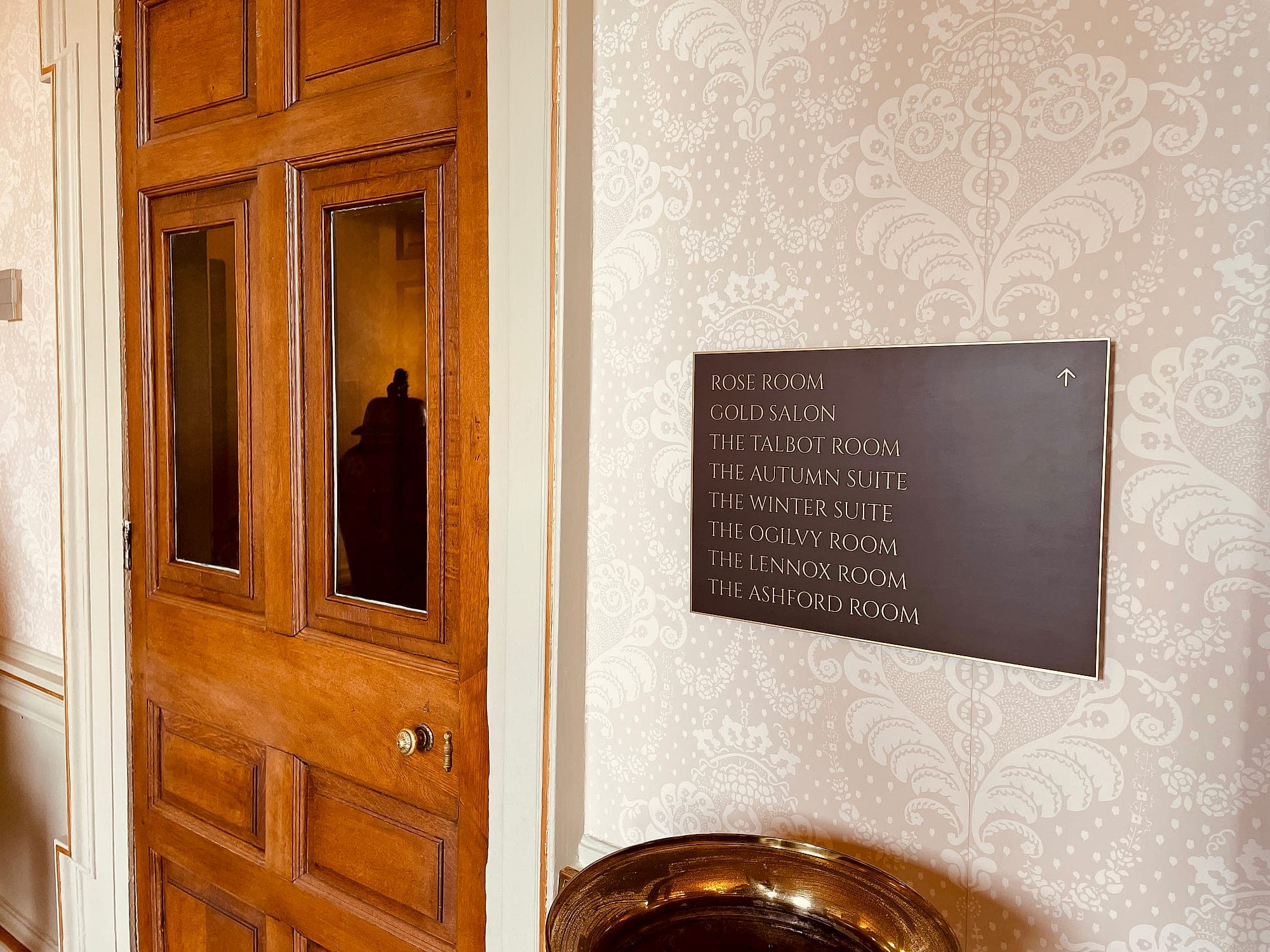 Signage installation in a heritage hotel: a dark directional panel with gold lettering fixed to patterned wallpaper beside a period panelled oak door at Carton House