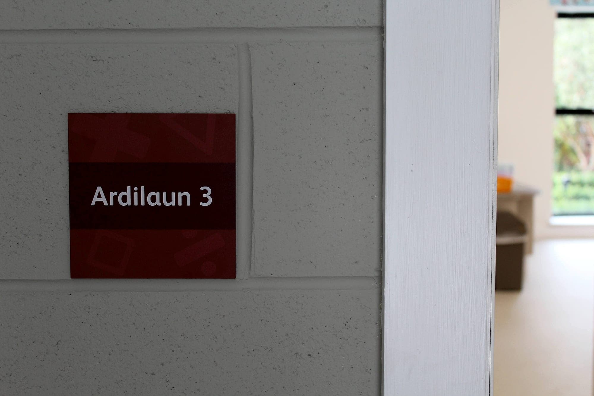 Signage installation on painted blockwork at Scoil Assaim: a maroon room identification panel fixed flat to a painted block wall, set within a single course and clear of the mortar joints
