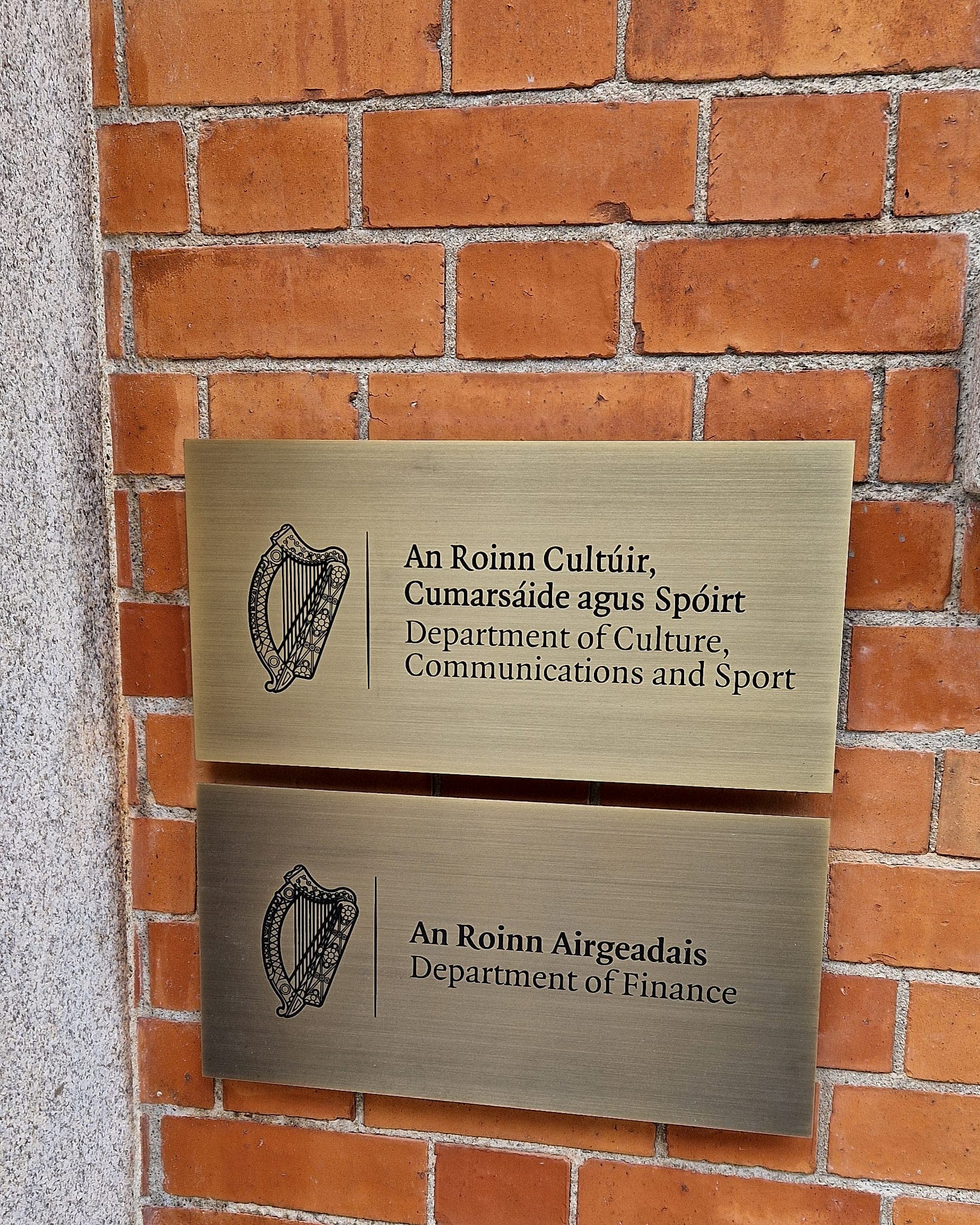 Two brass government department plaques at Leeson Lane, stood off a red brick facade beside a granite reveal, showing the barrel spacers and fixing positions set out during a signage site survey