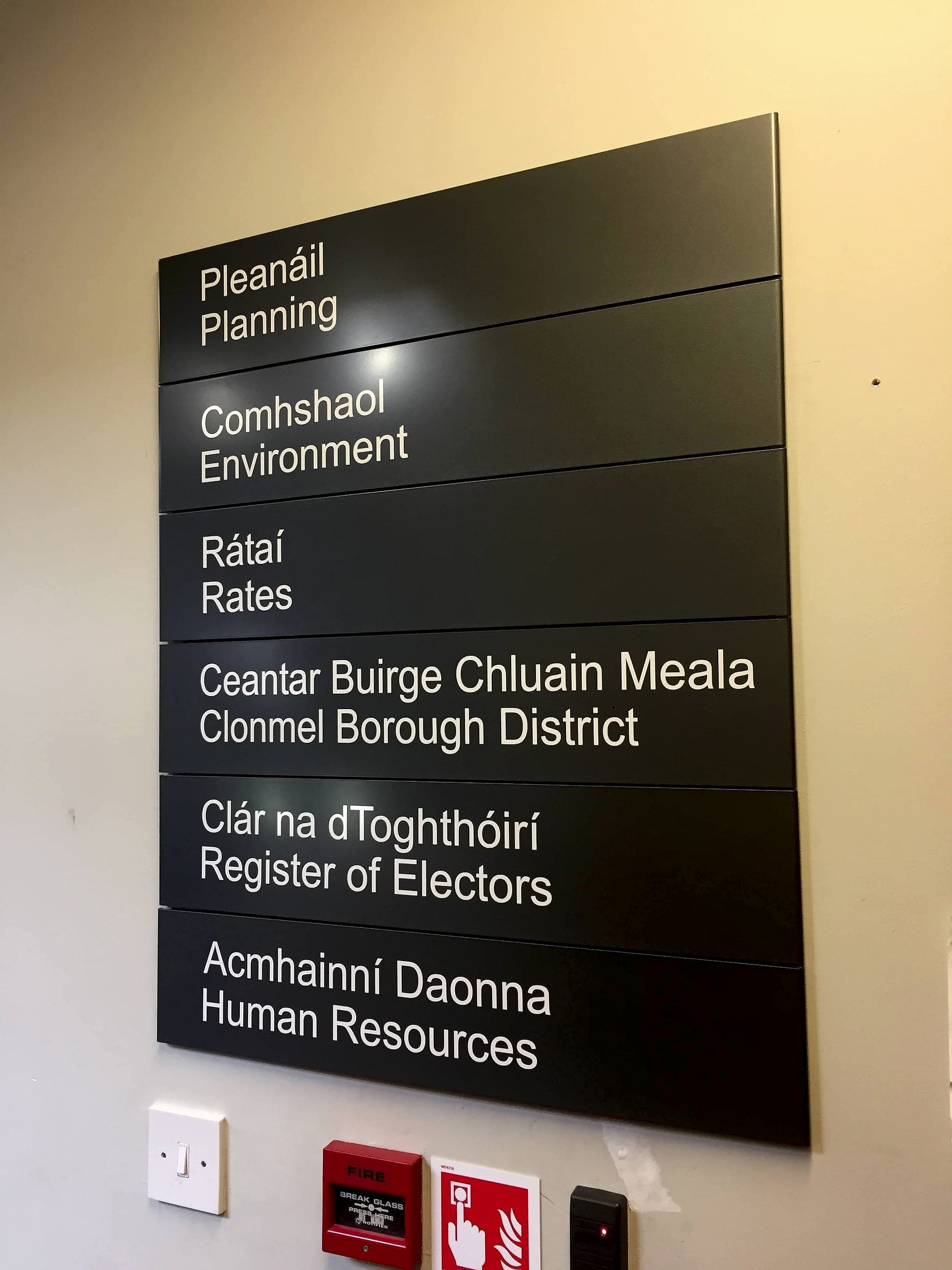 Bilingual Irish and English modular directory sign on a corridor wall in a Clonmel borough district office, mounted directly above a light switch, fire alarm call point, fire alarm sign and access-control reader