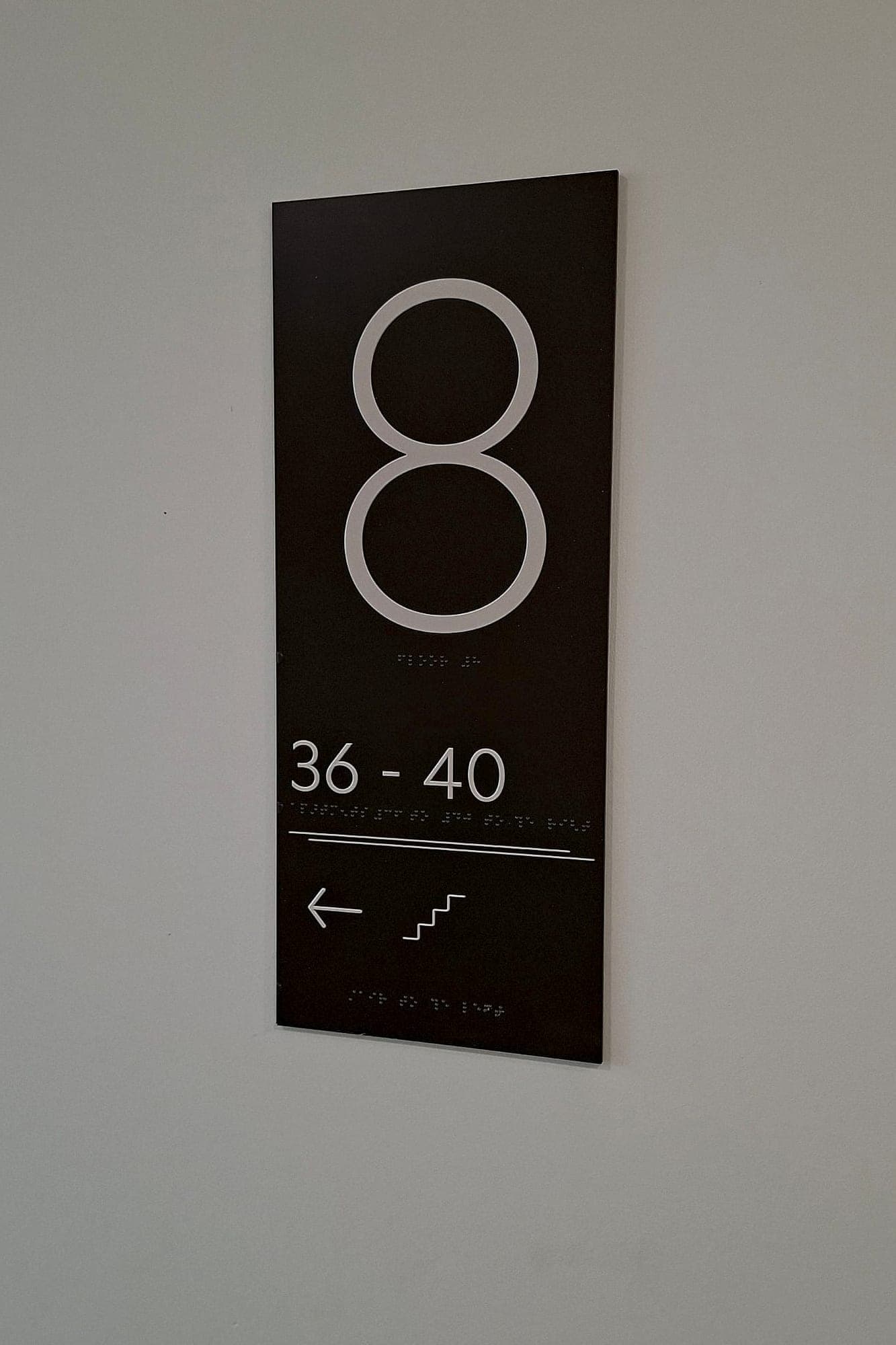 Residential stair-core sign with a large outlined level numeral, an apartment number range, a stair pictogram with a down arrow, and braille