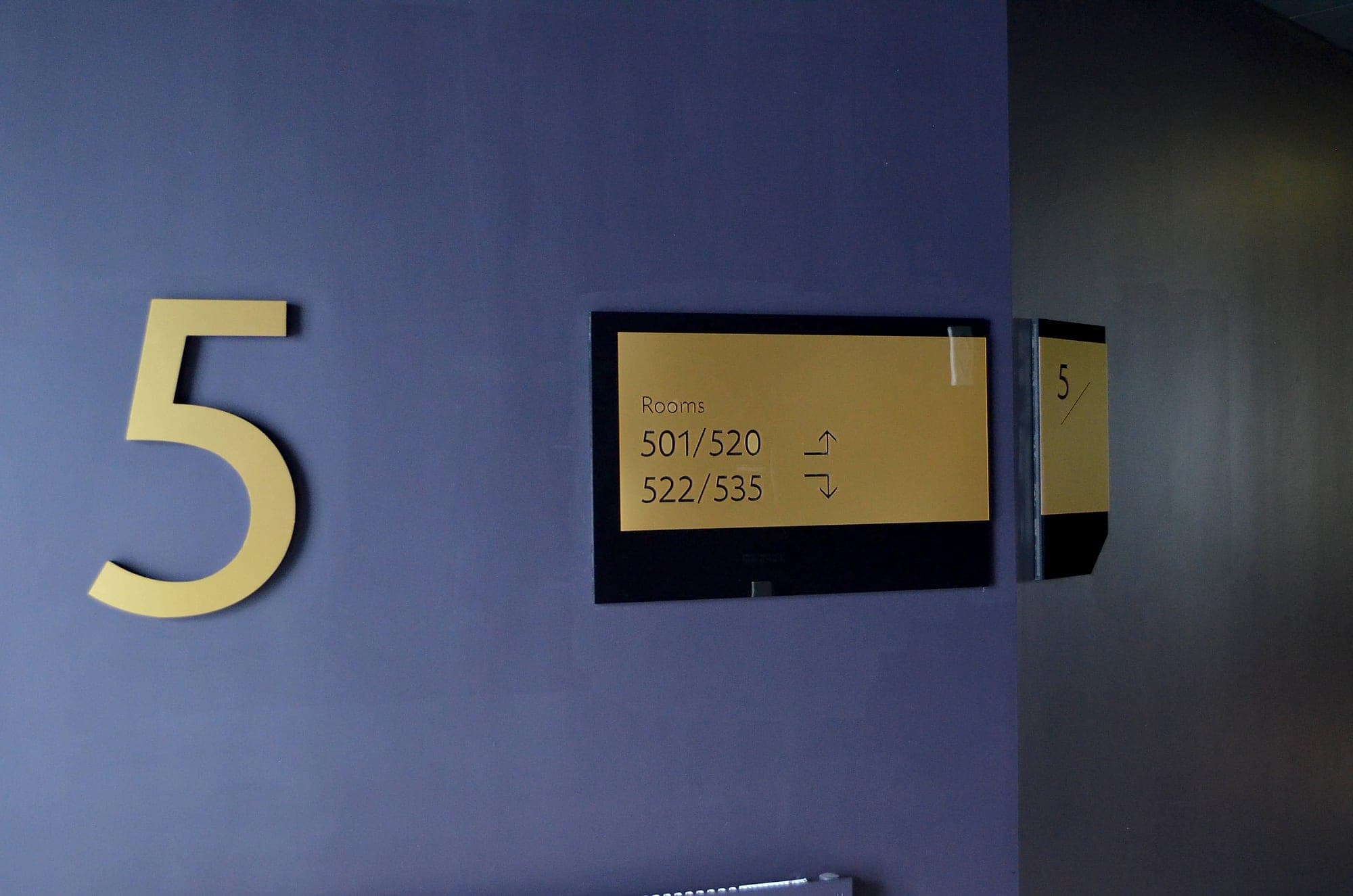 Digital vs static signage: a brushed gold floor numeral and a matching room-number range panel with directional arrows, mounted on a deep blue lift-lobby wall