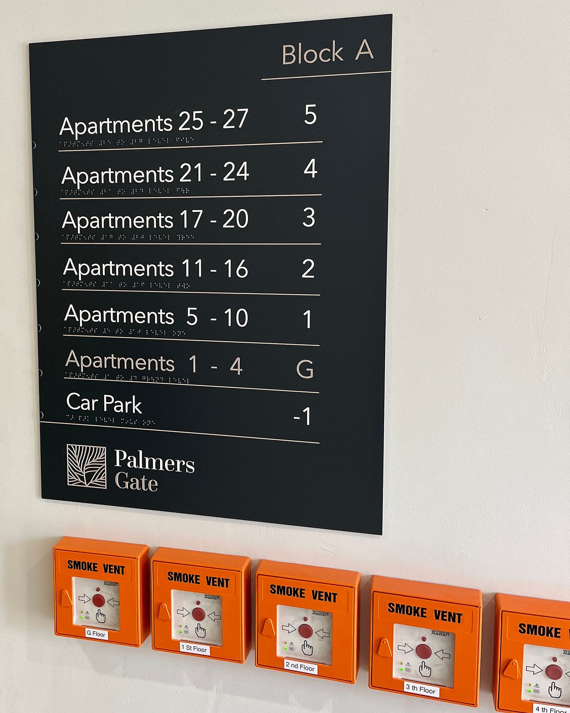 Statutory signage at handover: an apartment level directory with braille mounted above a row of orange smoke-vent control points labelled floor by floor