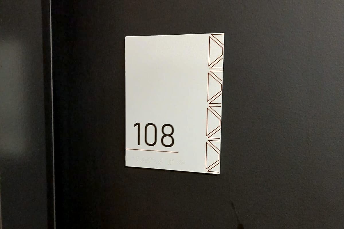 The door plate is the most-touched sign in the building. Student accommodation room door signage: a white door plate with the room number 108 in raised tactile numerals and braille beneath, on a dark door