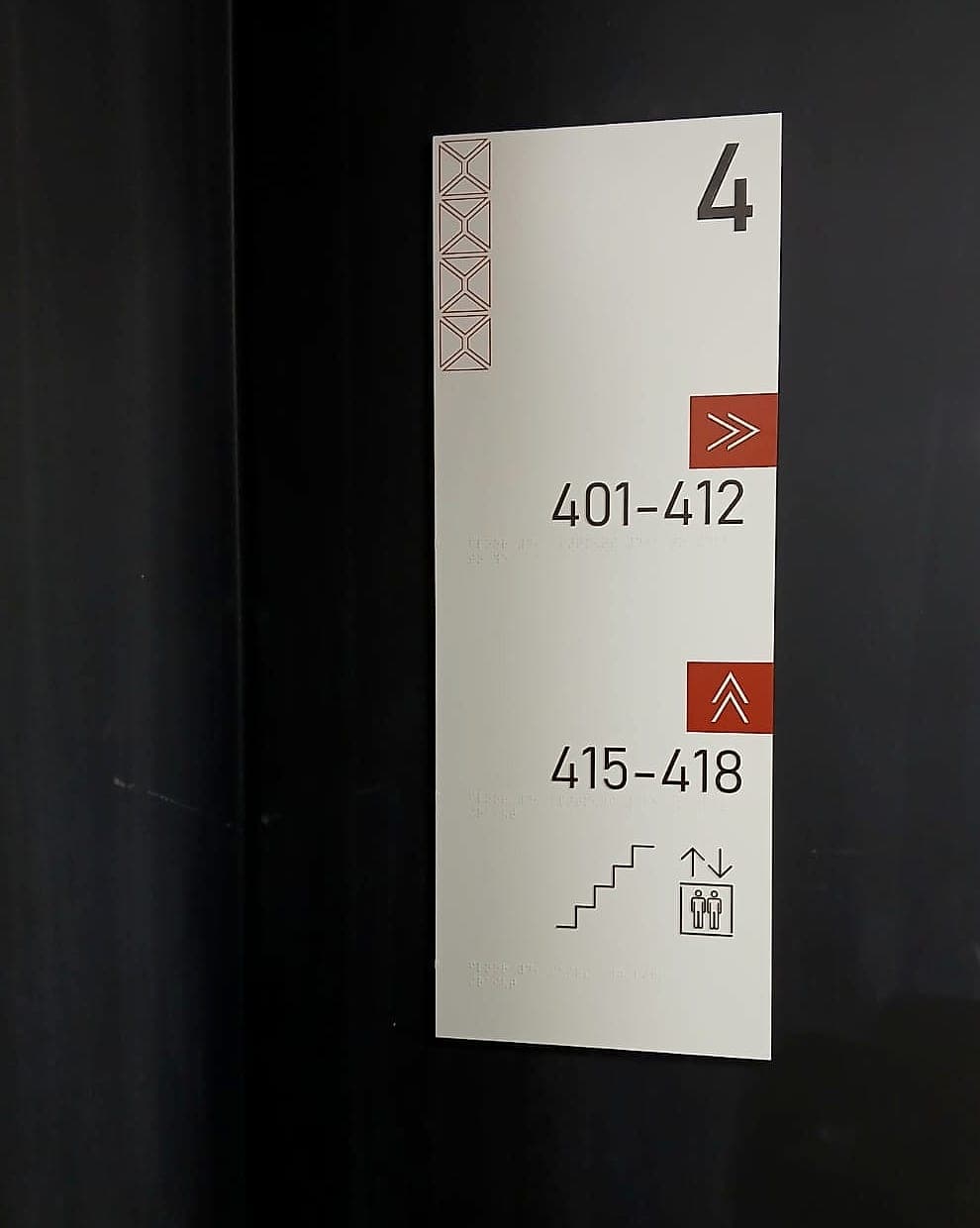 Student accommodation level directional sign on a dark corridor wall: floor number 4, room ranges 401-412 and 415-418 with arrows, stair and lift pictograms, and braille beneath each line