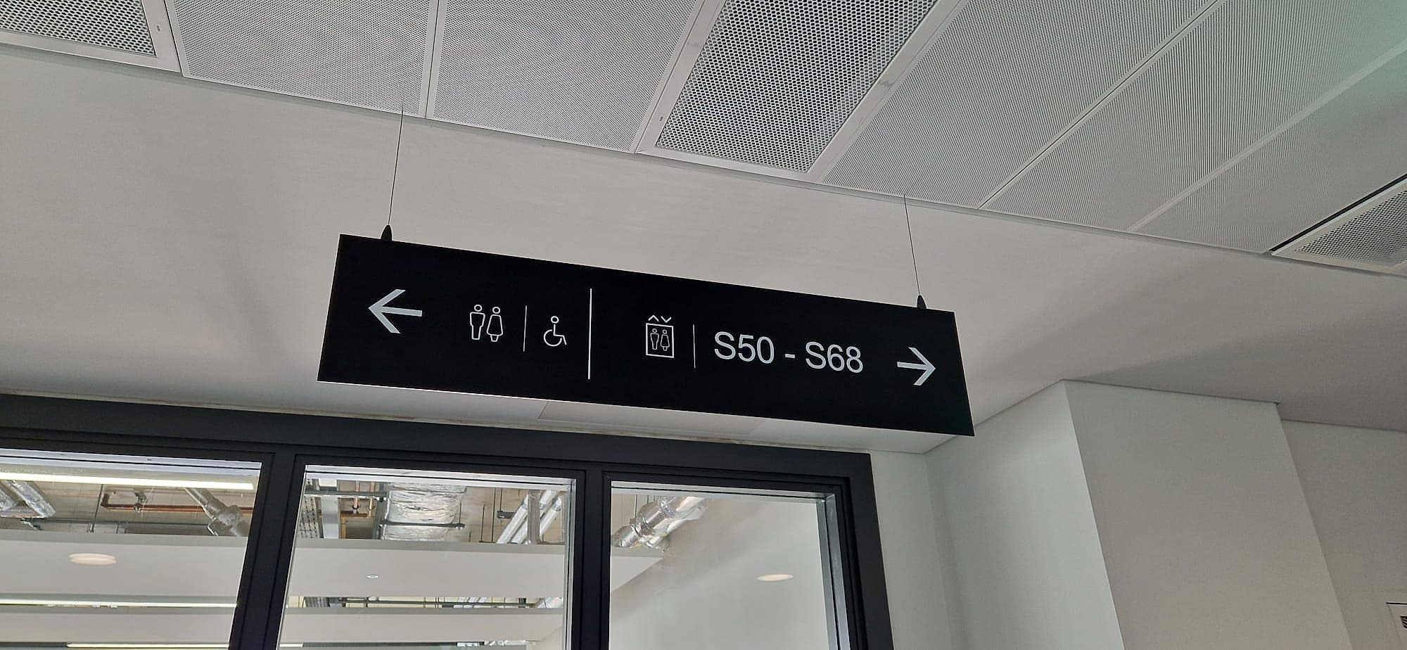Suspended directional wayfinding sign in a campus building in Athlone: an overhead panel with lift, toilet and accessibility pictograms and room ranges with arrows