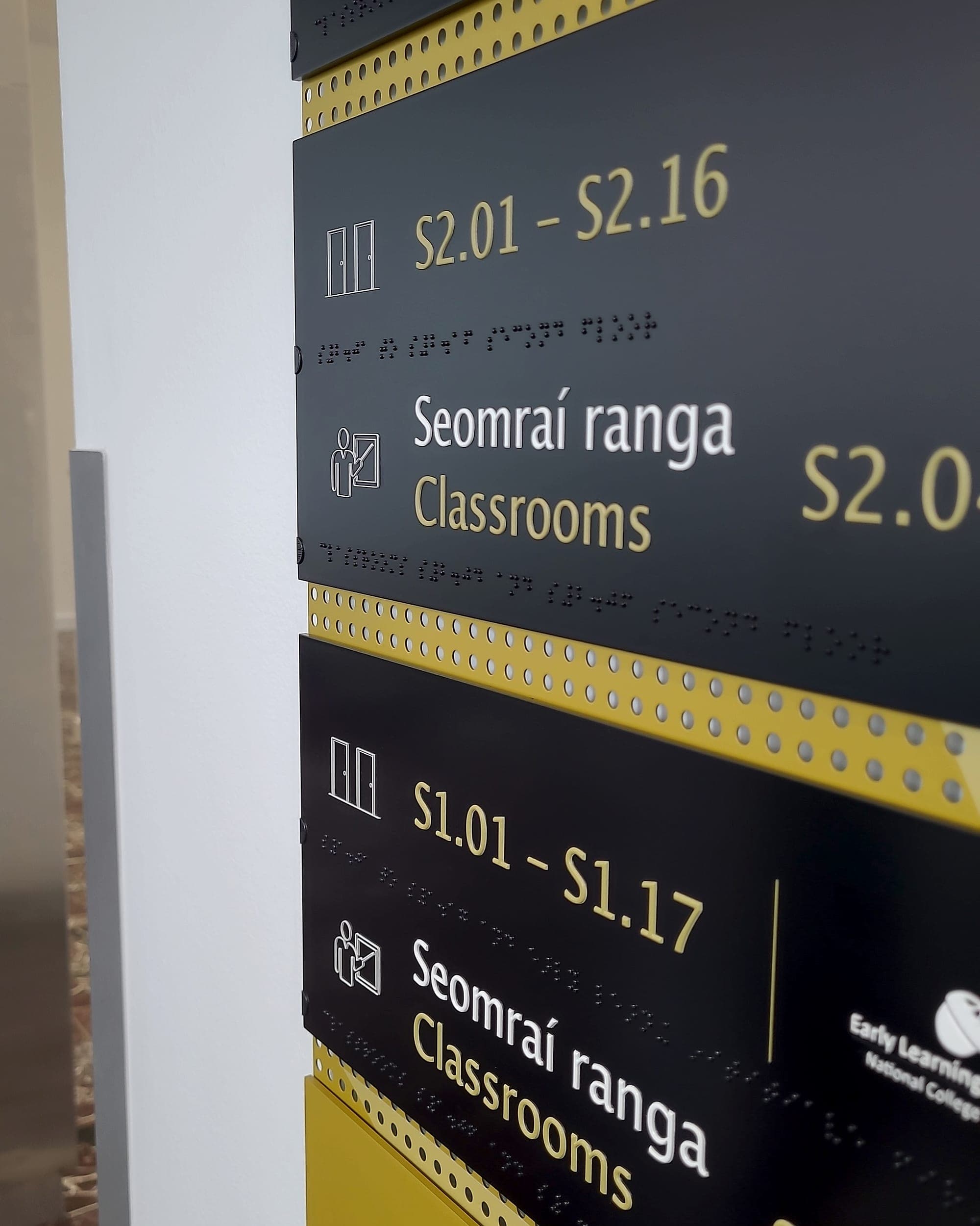 Close-up of bilingual tactile and braille directory signage at NCI Spencer Dock: angled black and yellow slats with raised Irish and English lettering, room-number ranges, pictograms and braille beneath each line