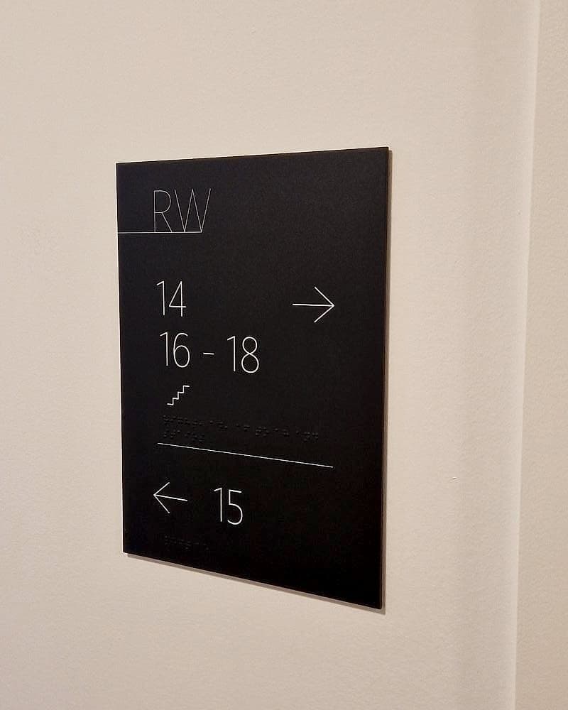 Tactile braille directional signage in an apartment corridor at Rathborne Wharf, Dublin: matt black panel with white flat numbers, stair pictogram and braille lines, mounted at reading height on a corridor wall