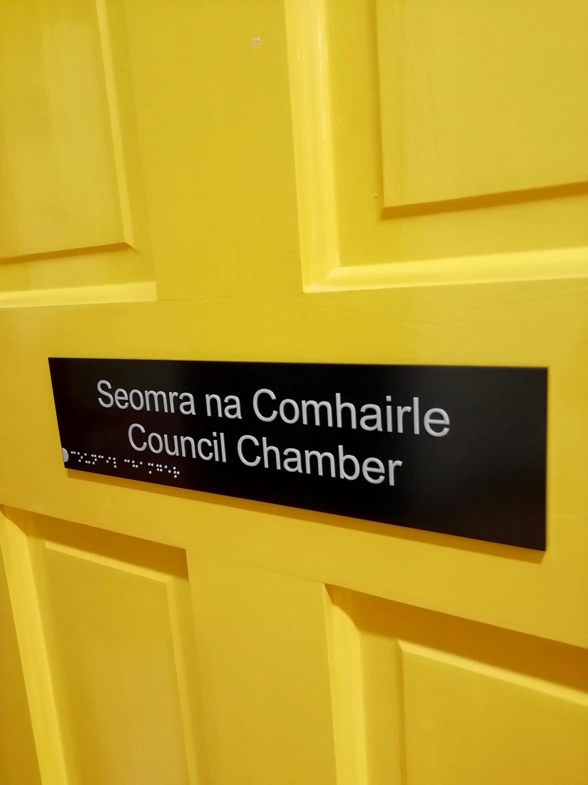 A council chamber door with a high-contrast bilingual tactile door sign: raised white mixed-case Irish and English lettering with braille on a black panel, mounted at hand height on a bright yellow council chamber door