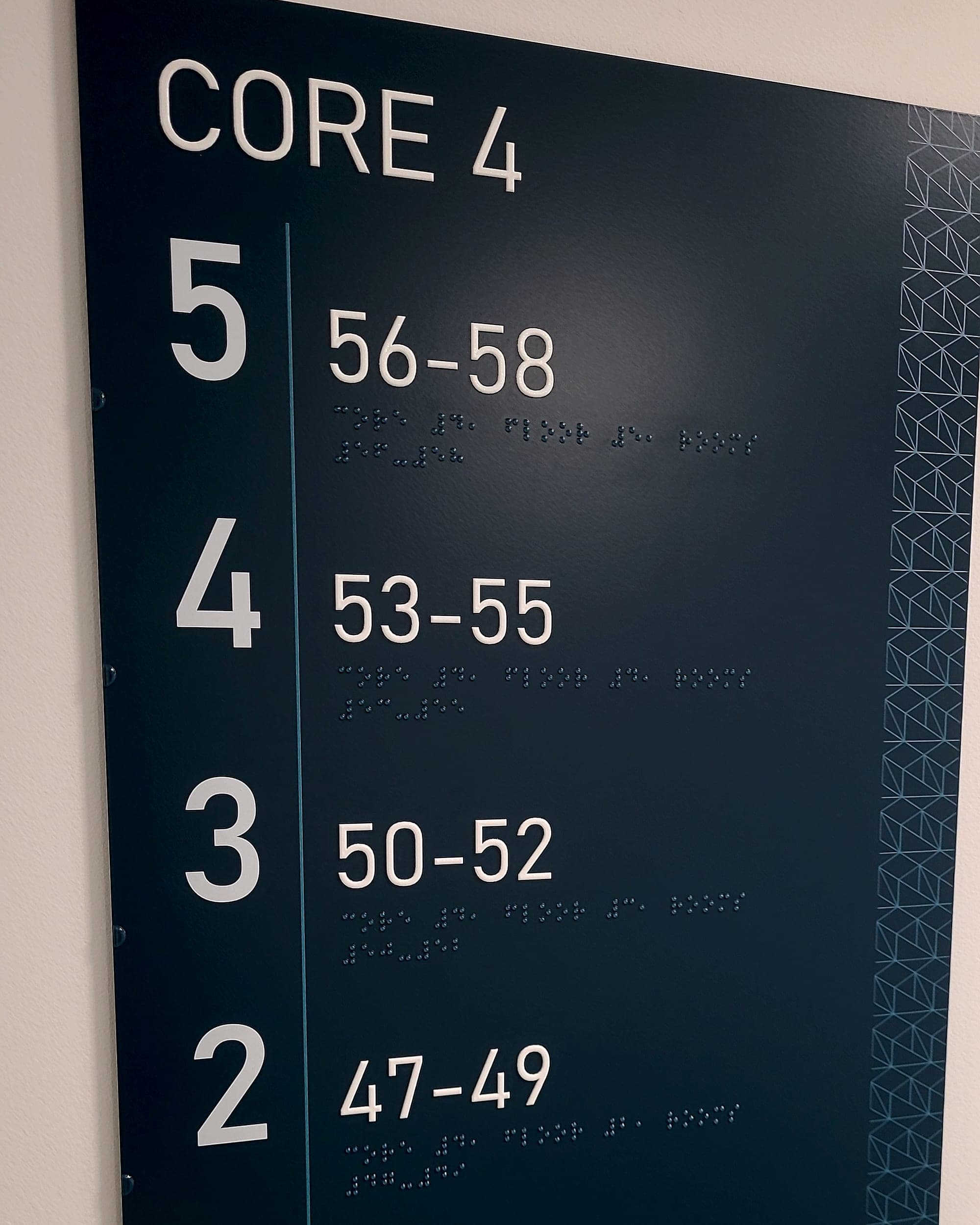 A Dublin apartment scheme: wall-mounted residential core directory in dark teal, listing each level against its apartment-number range in raised tactile numerals with braille beneath every line