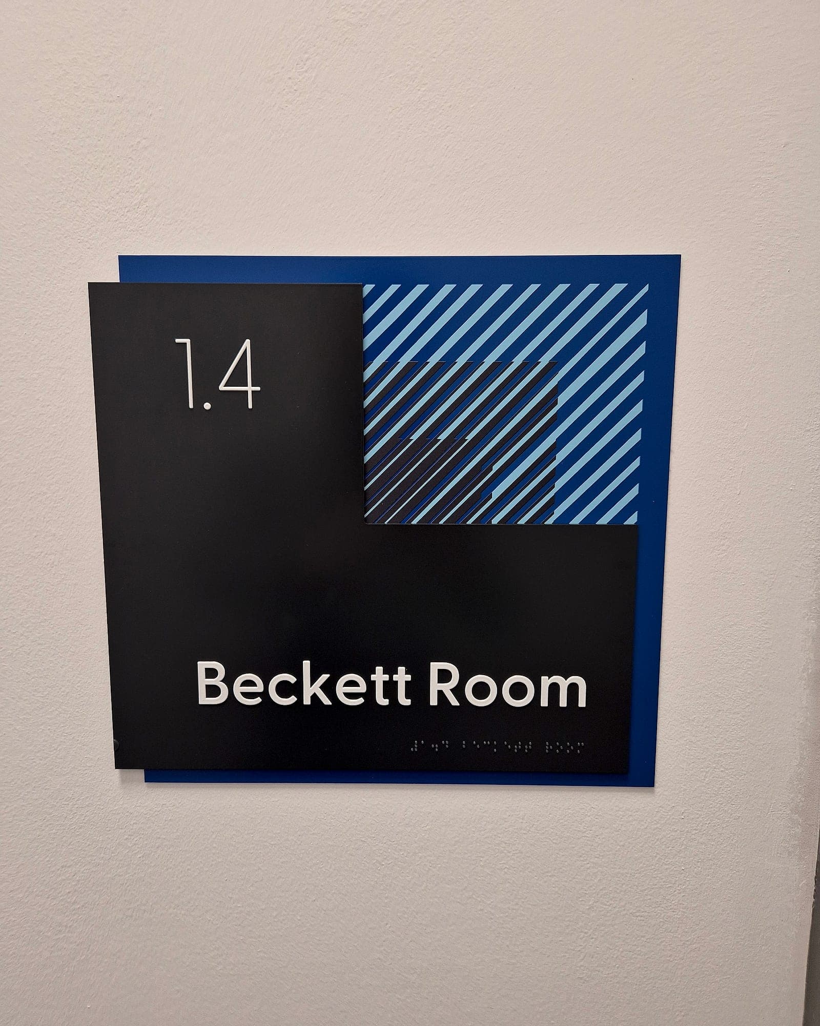Tactile room sign at CCT College with raised lettering and braille reading 1.4 Beckett Room, layered black panel on blue with a striped graphic block