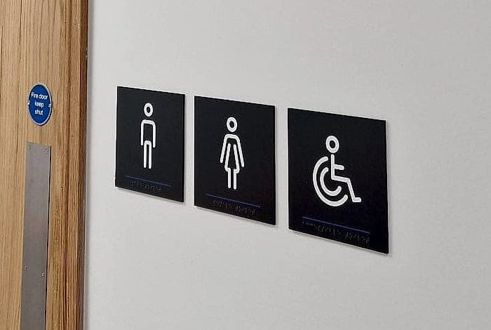 Tactile braille toilet signage at CCT College Dublin: three dark panels with raised white male, female and wheelchair pictograms, each with a braille line beneath, mounted in a row at reading height beside a timber door