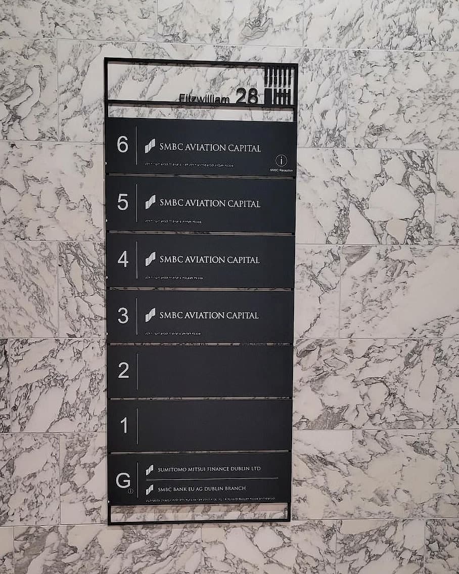 Multi-let office building directory for SMBC built as changeable slats: four floors carrying one tenant's name, two blank slats for vacant levels, two ground-floor occupiers, with braille under each line