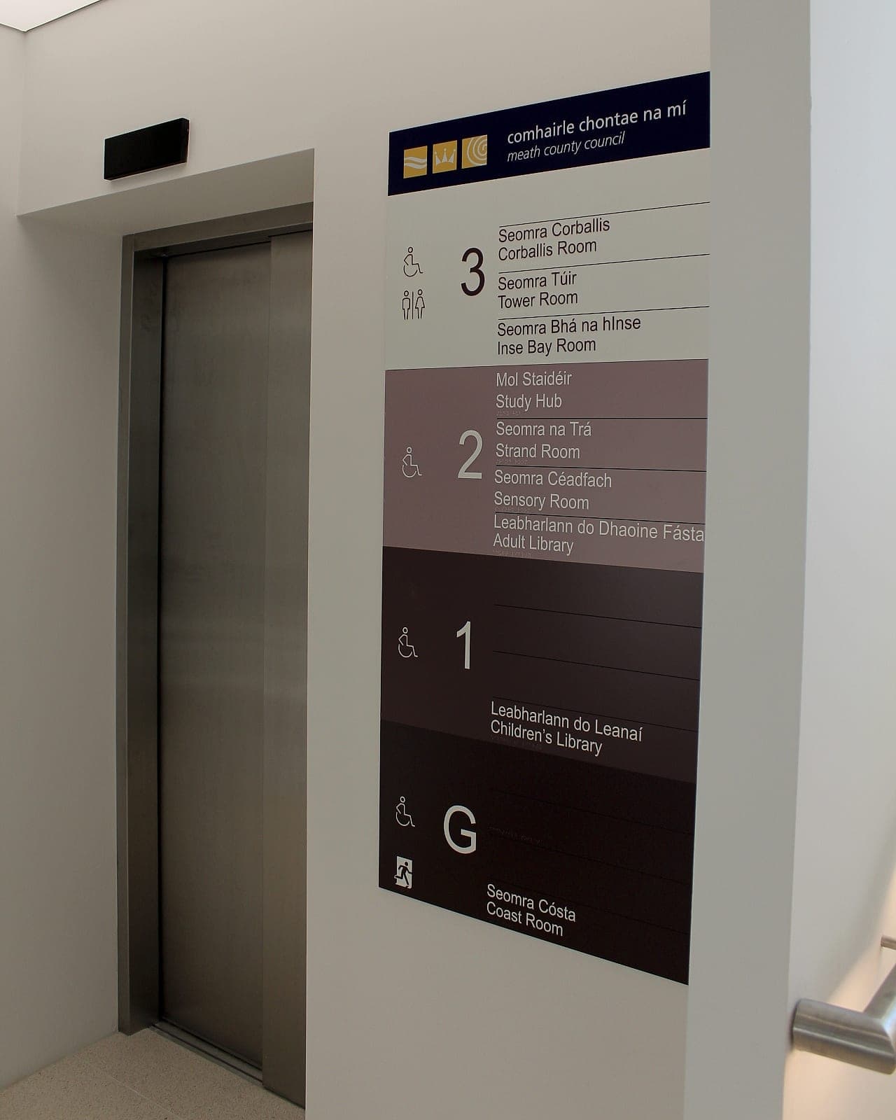 Bettystown Library, bilingual lift-lobby directory for Meath County Council: a tonal band panel where dark text on the pale band and white text on the dark bands read strongly while the mid-tone band recedes