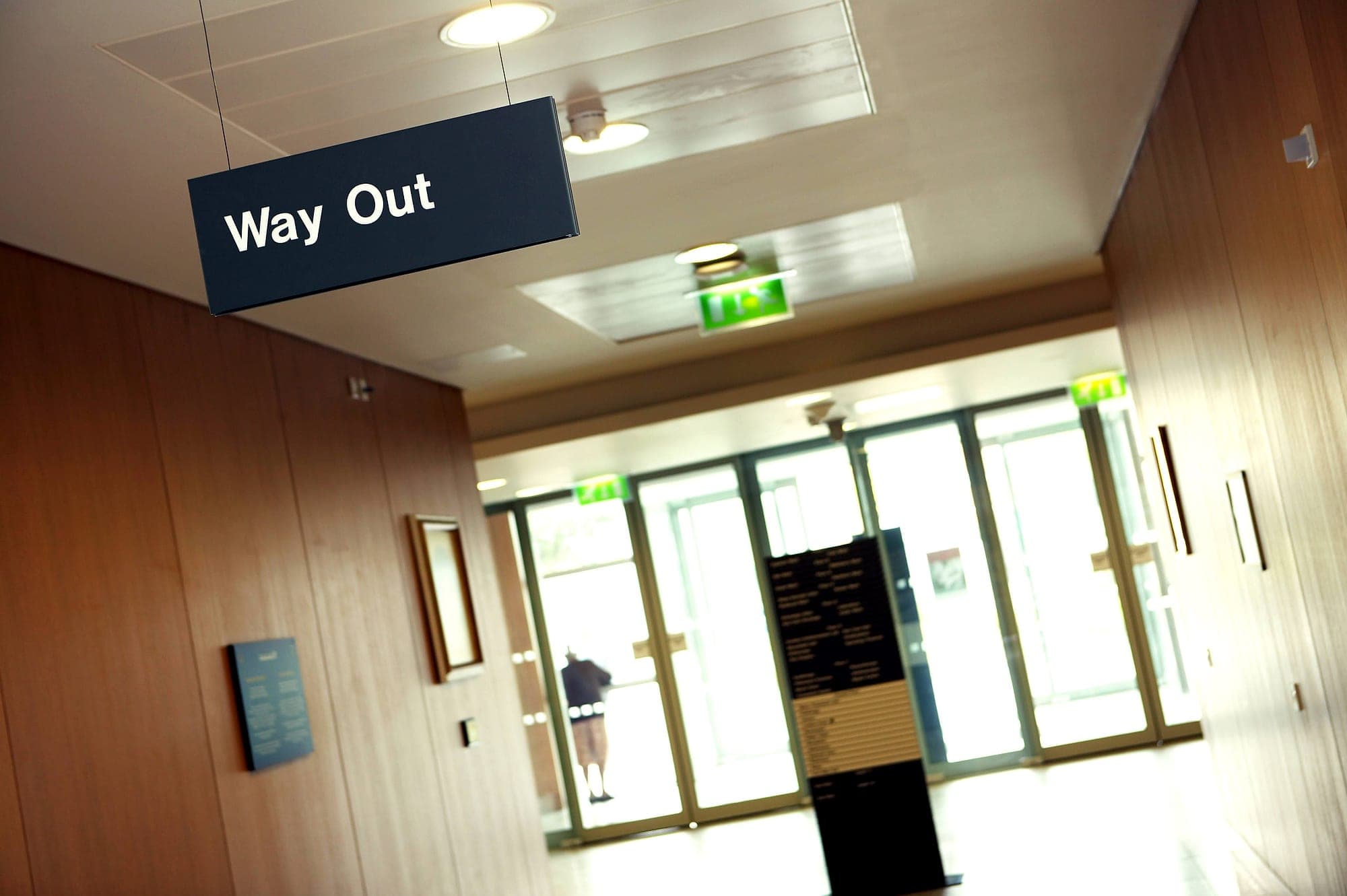 St Vincent's Private Hospital: navy suspended Way Out wayfinding sign in a timber-lined lobby, with green maintained emergency exit signs above the glazed final exit doors beyond