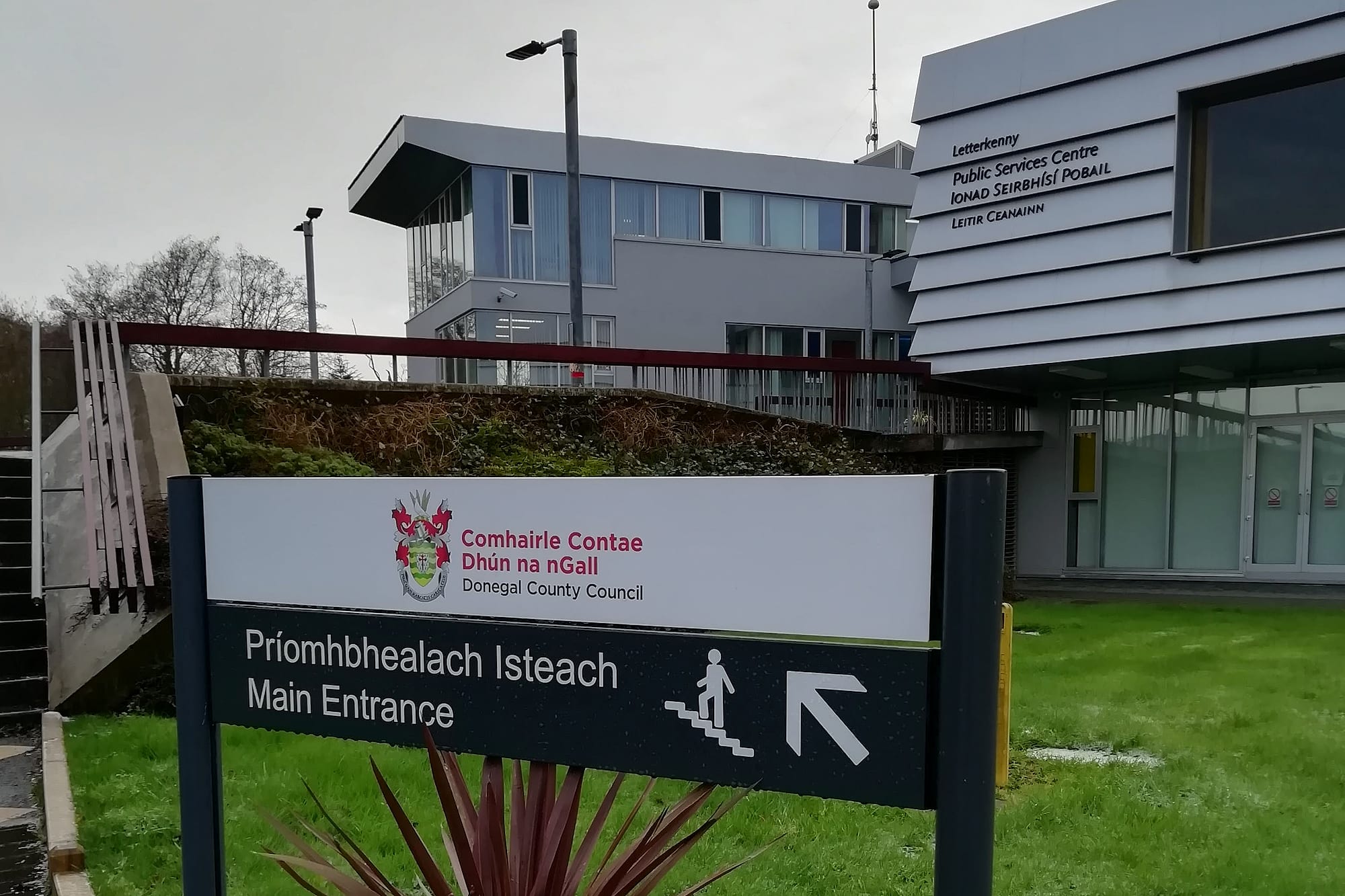 Council signage at Donegal County Council's Letterkenny Public Services Centre, a bilingual Irish and English post-and-panel main entrance sign with the county crest, and the building's own bilingual identification lettering on the facade behind