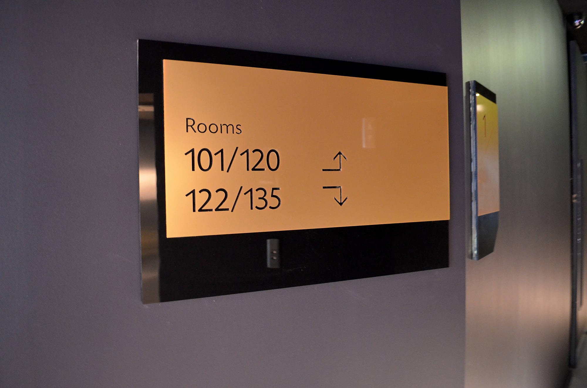 Lift-lobby directional sign at The Marker Hotel showing room number ranges 101/120 and 122/135 with arrows on a bronze panel