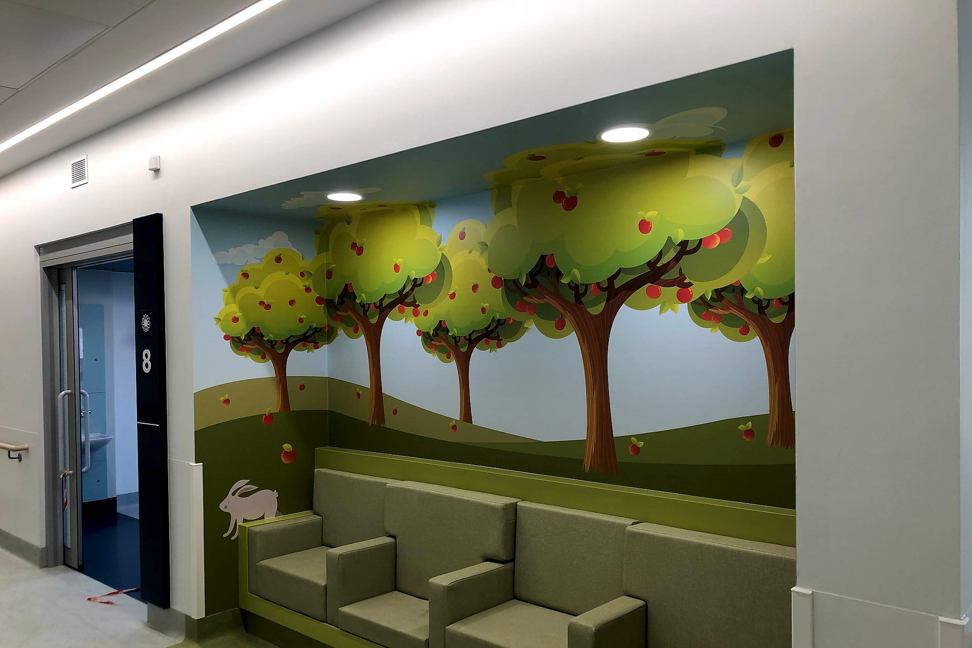 Apple-orchard wall mural running the full height of a corridor seating bay at the National Rehabilitation Hospital, beside a door pier carrying the numeral 8 and a flower symbol